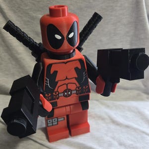 May include: A red and black Deadpool Lego figure with black weapons. The figure has a red head with white eyes and a black mask. The torso is red with black detailing. The figure is holding two black guns.