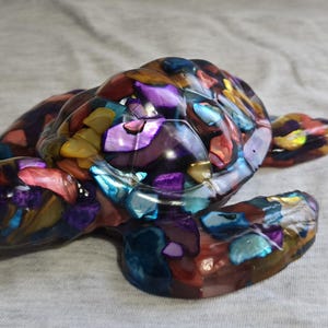 May include: A colorful sea turtle figurine made of iridescent mosaic tiles. The turtle is in a resting position with its head turned to the side. The mosaic tiles are in shades of blue, green, purple, pink, and gold.
