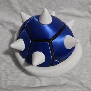 May include: A blue and white novelty item resembling a spiky turtle shell. The shell is a vibrant blue with black accents and white spikes. The base is white, creating a striking contrast. This item is likely a collectible or decorative piece.