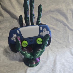May include: A green and purple 3D printed hand holding a blue PlayStation 5 controller with green buttons. The controller is resting in the palm of the hand, with the thumbsticks facing forward. The hand is posed in a 'stop' gesture, with the index finger extended and the other fingers curled.