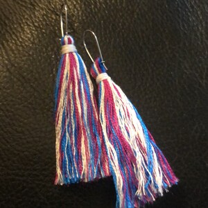 Op de afbeelding: Een paar oorhangers met blauwe, witte en roze kwastjes. De oorhangers zijn gemaakt van zilverkleurig metaal.
