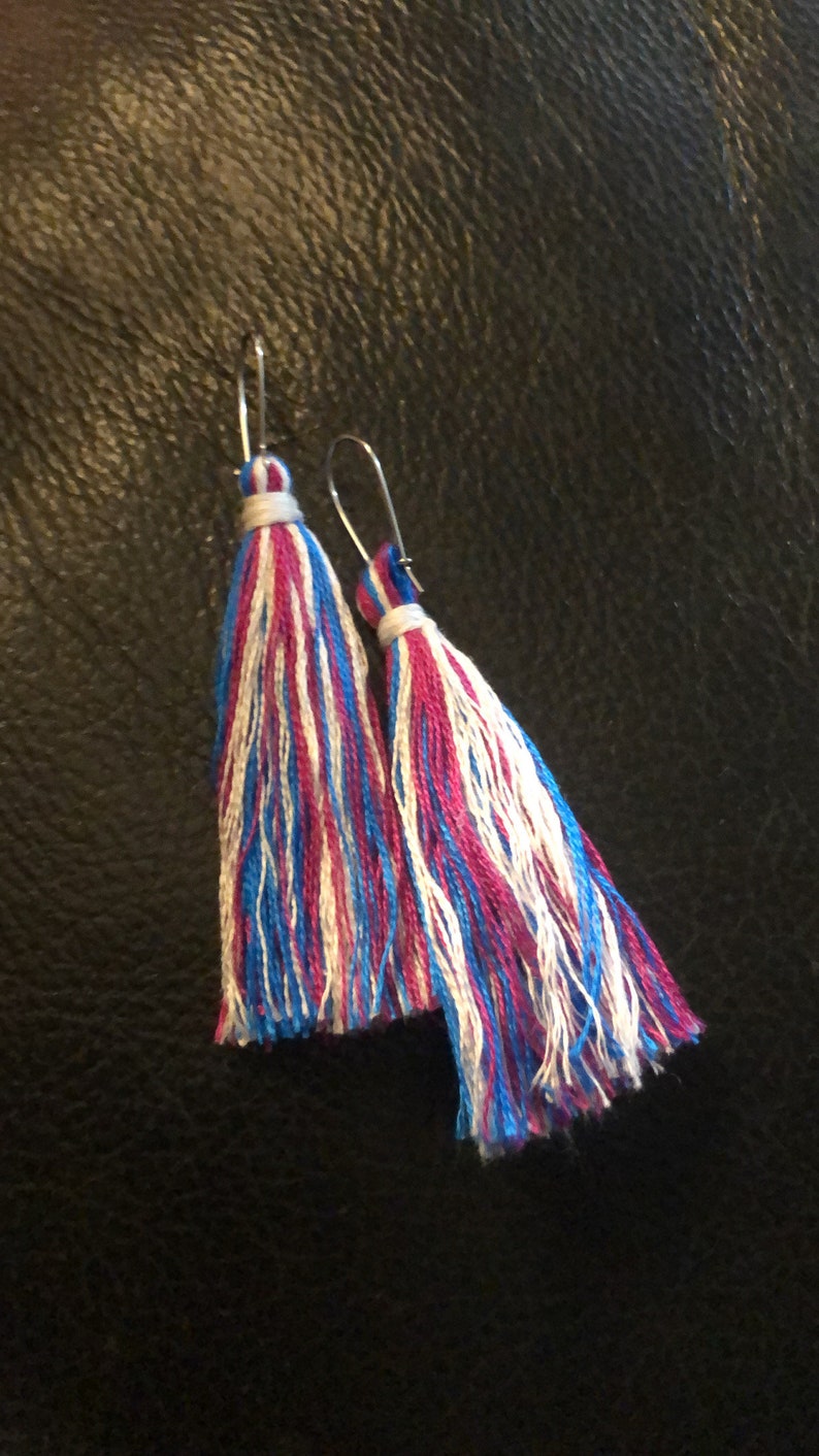 Op de afbeelding: Een paar oorhangers met blauwe, witte en roze kwastjes. De oorhangers zijn gemaakt van zilverkleurig metaal.