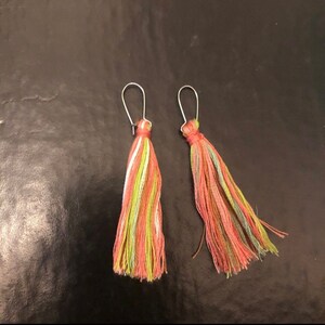 Op de afbeelding: Een paar oorbellen met kleurrijke kwastjes. De kwastjes zijn gemaakt van garen in tinten roze, groen en wit. De oorbellen zijn zilver.