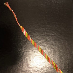 May include: Handmade friendship bracelet featuring a woven pattern of orange, green, brown, and white threads. The bracelet has a knotted end and a loop for closure, showcasing a vibrant and colorful design. This accessory is perfect for casual wear.