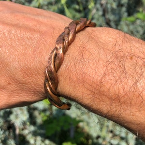 May include: A copper-colored, braided cuff bracelet. The bracelet has a textured, hammered appearance and is worn on a wrist. The design features multiple strands twisted together, creating a unique and rustic look. The bracelet is a piece of jewelry.