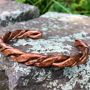 May include: A handcrafted copper cuff bracelet with a twisted rope design. The open cuff features a hammered texture and a warm, reddish-brown color. This artisan jewelry piece is displayed on a textured stone surface.