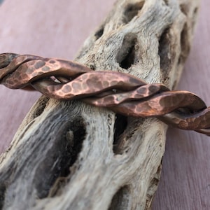 May include: A handcrafted copper bracelet with a twisted design. The bracelet has a hammered texture and a warm, reddish-brown color. The bracelet is displayed on a piece of driftwood.