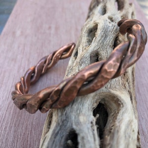 May include: A copper-colored, twisted metal bracelet. The cuff bracelet has a hammered texture and a braided design. The jewelry is displayed on a piece of driftwood, showcasing the bracelet's rustic appeal.