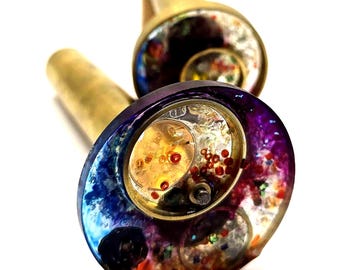 May include: Two decorative bottle stoppers with brass stems and colorful, swirling resin tops. The resin is a mix of blues, purples, oranges, and reds, with embedded small, round, red and clear elements. The stoppers are set against a white background.