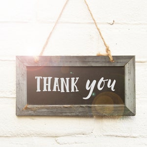 May include: A rectangular wooden sign with the words "THANK you" in white lettering. The sign is framed in gray wood and hangs from a natural twine rope against a white brick wall.