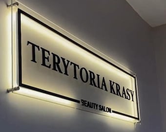 May include: Illuminated sign for a beauty salon, mounted on a wall. The sign features the name "TERYTORIA KRASY" in bold, black letters, with "BEAUTY SALON" below. The sign has a black border and a warm, glowing light.