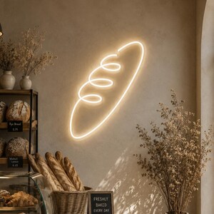 May include: A warm white neon sign shaped like a loaf of bread mounted on a beige wall above a bakery counter. The counter features a glass display case filled with various baked goods, a wicker basket of baguettes, and a small sign reading Freshly Baked Every Day.