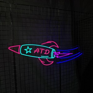 May include: A neon sign in the shape of a rocket ship, with a teal body and pink accents. The rocket has the letters "ATD" and a star on its side. The exhaust is blue and purple.