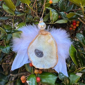 May include: A handcrafted angel ornament made from a geode slice with gold trim, white feathers, and a pearl bead head. The ornament hangs from a clear string against a backdrop of green leaves and red berries.