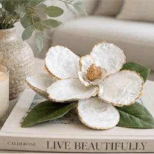 May include: A decorative white and gold magnolia flower sculpture with green leaves, resting on a book with the words "LIVE BEAUTIFULLY". The flower has a gold center and gold-trimmed petals. A vase with greenery and a candle are in the background.