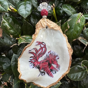 May include: An oyster shell ornament with a gold-painted edge and a red and black Gamecock logo. The ornament is suspended by a beaded strand with red, black, and clear beads. The background is a lush green plant.