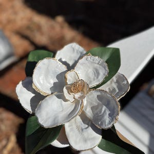 May include: A large, handcrafted white flower with gold-painted edges and a textured center. The flower is surrounded by green leaves. The flower is likely a decorative item or a piece of art.