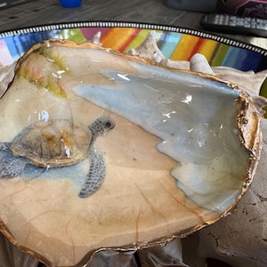 May include: A decorative oyster shell dish featuring a painted sea turtle on a sandy beach scene. The shell has a gold-colored rim and is set against a colorful background. The turtle is depicted in shades of brown, green, and blue.