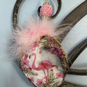 May include: A decorative seashell ornament featuring a pink flamingo design. The shell is adorned with pink flowers and trimmed with gold. Pink feathers and a beaded accent add to the charm. A unique, handcrafted item.