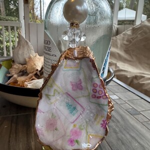 May include: A decorative bottle stopper featuring a white oyster shell with a gold-colored edge. The shell is decorated with a floral and butterfly pattern. The stopper has a pearl and crystal bead design.
