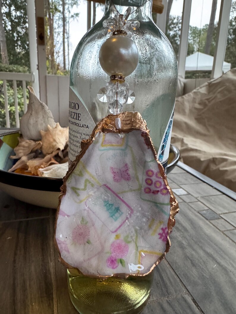 May include: A decorative bottle stopper featuring a white oyster shell with a gold-colored edge. The shell is decorated with a floral and butterfly pattern. The stopper has a pearl and crystal bead design.