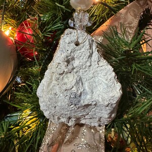 May include: A unique, handmade Christmas tree ornament. The ornament is a rough, textured, off-white stone shape, suspended by a string and adorned with a pearl bead and crystal accents. The background features a decorated Christmas tree with gold ribbon and lights.