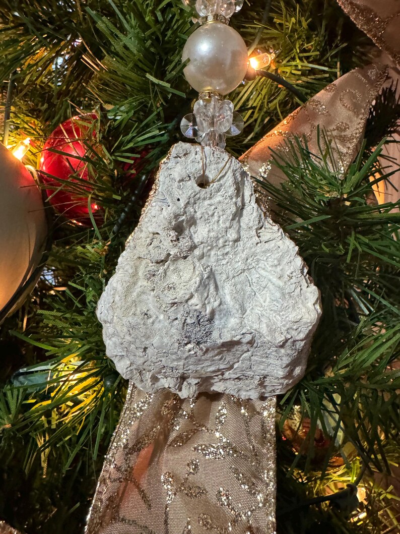 May include: A unique, handmade Christmas tree ornament. The ornament is a rough, textured, off-white stone shape, suspended by a string and adorned with a pearl bead and crystal accents. The background features a decorated Christmas tree with gold ribbon and lights.