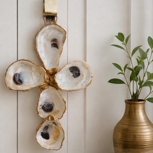 May include: A decorative wall hanging made from five oyster shells, arranged in a cross shape. The shells have a gold-painted trim and are attached to a brass-colored metal clip with a natural fiber cord. The shells are white with dark markings.