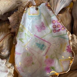 May include: A decorative oyster shell with a gold-painted rim. The shell interior features a floral and butterfly design with the word "JOKER" visible. The shell is surrounded by other shells.