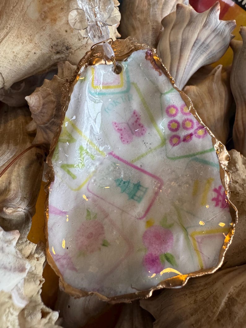 May include: A decorative oyster shell with a gold-painted rim. The shell interior features a floral and butterfly design with the word "JOKER" visible. The shell is surrounded by other shells.