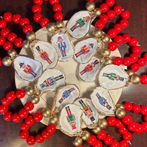 May include: A set of 12 nutcracker ornaments, each painted on a white oyster shell with gold trim. The ornaments are arranged in a circle on a white plate, surrounded by red beaded napkin rings.