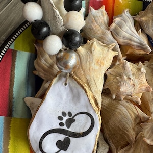 May include: A beaded garland with black, white, and tan wooden beads. A white oyster shell pendant with a black paw print, infinity symbol, and heart design. The pendant is surrounded by seashells.