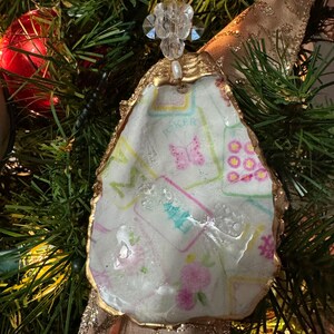 May include: An oyster shell Christmas ornament with a gold-painted edge. The shell is decorated with a floral and butterfly pattern in pink, blue, and green. The ornament is attached to a gold ribbon and a pearl-topped embellishment.
