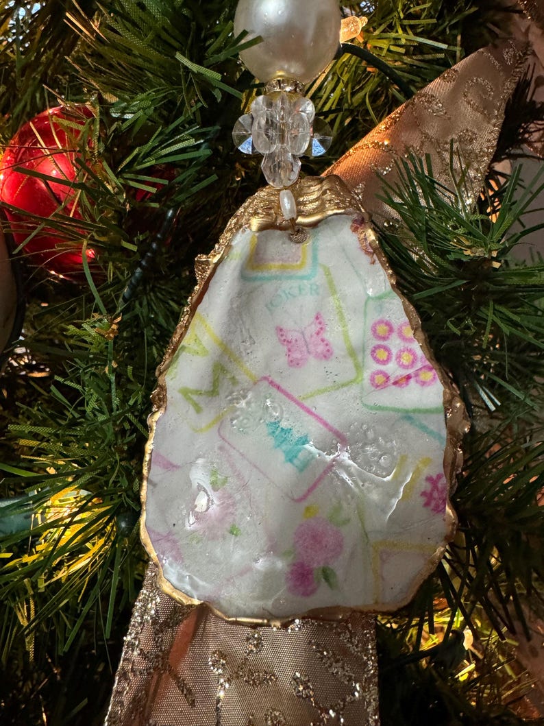 May include: An oyster shell Christmas ornament with a gold-painted edge. The shell is decorated with a floral and butterfly pattern in pink, blue, and green. The ornament is attached to a gold ribbon and a pearl-topped embellishment.