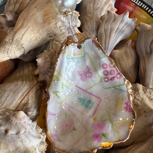 May include: A handcrafted oyster shell pendant with a gold-painted edge. The shell is decorated with a floral and geometric pattern in pink, green, and blue. The pendant is strung on a clear beaded strand with a large pearl accent.