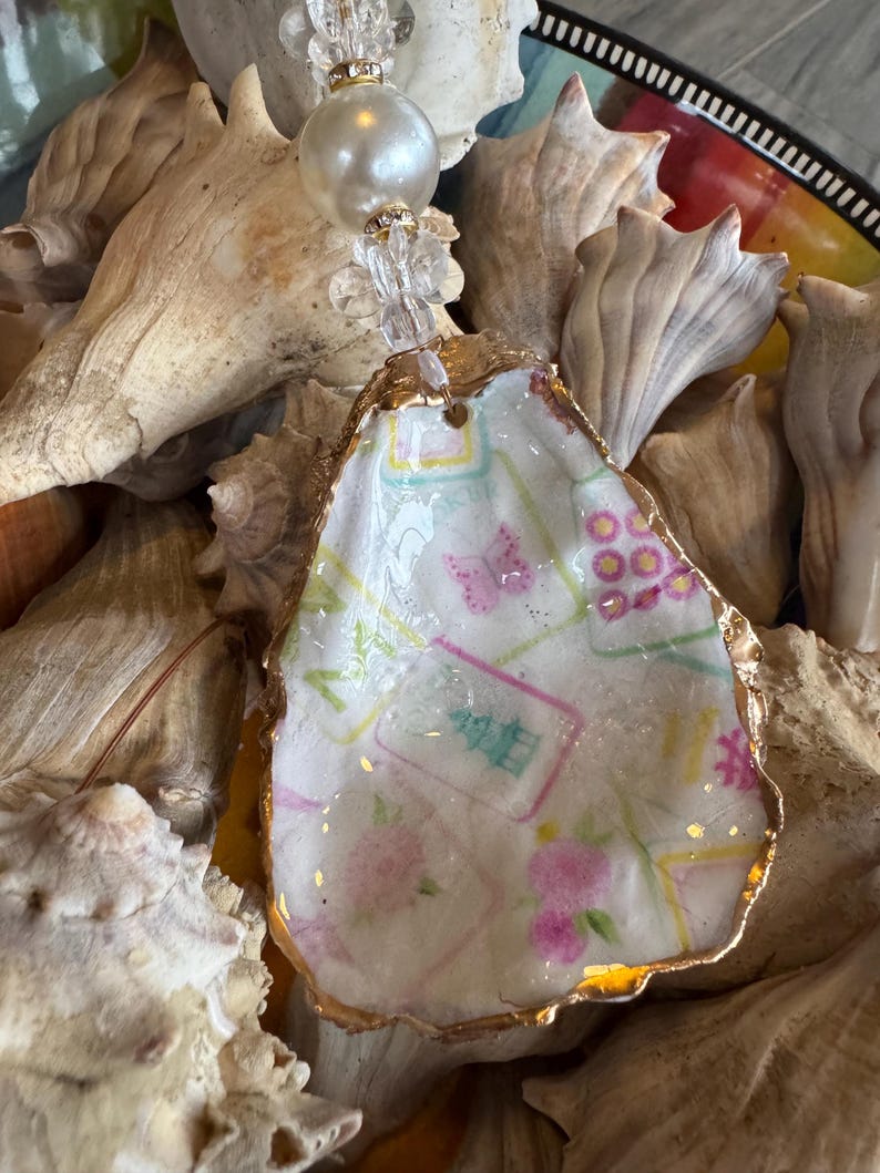 May include: A handcrafted oyster shell pendant with a gold-painted edge. The shell is decorated with a floral and geometric pattern in pink, green, and blue. The pendant is strung on a clear beaded strand with a large pearl accent.