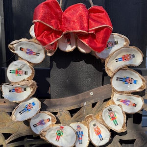 May include: A festive Christmas wreath made from oyster shells, each adorned with a colorful nutcracker image. A large red bow with gold trim sits at the top. The wreath is suspended by a natural twine loop, perfect for holiday decor.