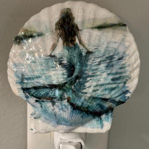 May include: A decorative night light featuring a seashell design. The seashell is painted with a mermaid in a blue dress in the water. The night light has a white base with an on/off switch.