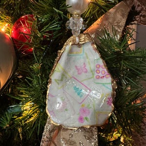 May include: A handmade Christmas ornament made from an oyster shell. The shell is painted with a pastel floral and butterfly design, edged with gold paint. The ornament is suspended by a beaded strand and hangs on a Christmas tree with gold ribbon.