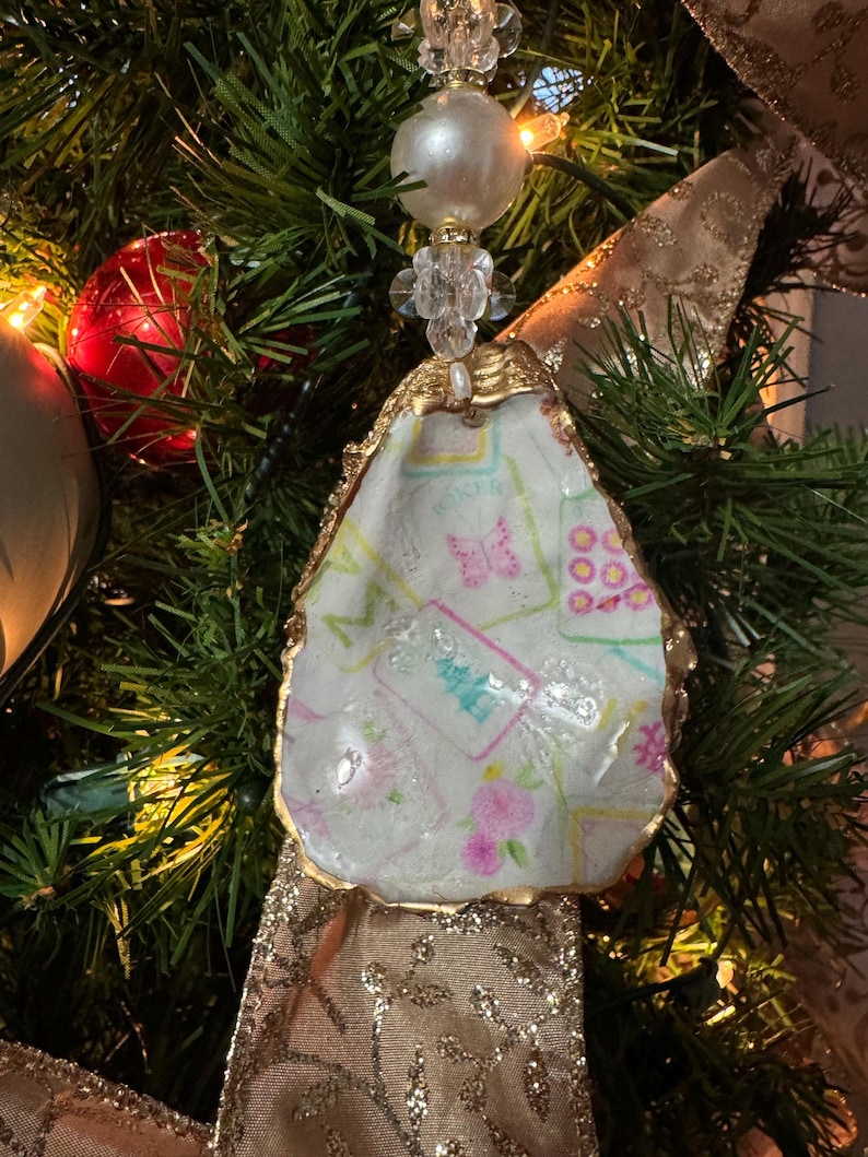 May include: A handmade Christmas ornament made from an oyster shell. The shell is painted with a pastel floral and butterfly design, edged with gold paint. The ornament is suspended by a beaded strand and hangs on a Christmas tree with gold ribbon.