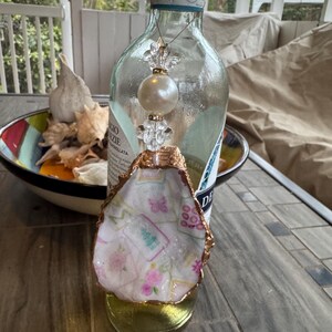 May include: An oyster shell ornament with a gold-painted edge, featuring a floral pattern in pink and green. A clear glass bottle with a decorative beaded charm hangs above the shell. The ornament sits on a gold-colored base.