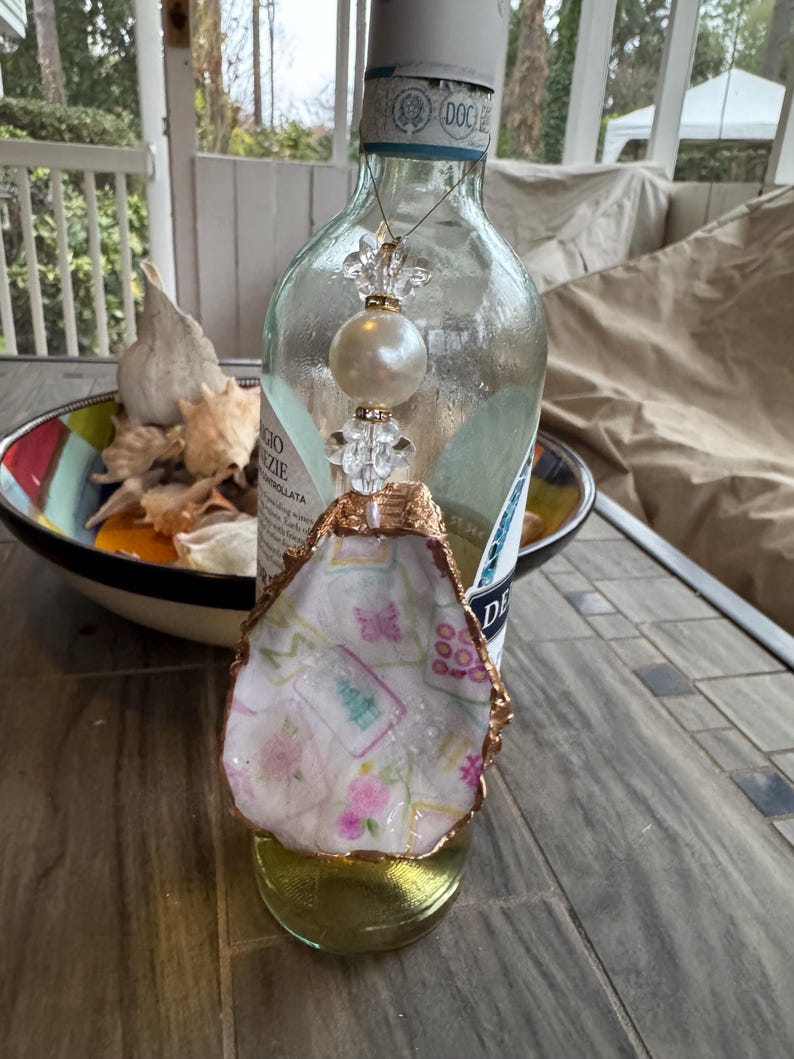 May include: An oyster shell ornament with a gold-painted edge, featuring a floral pattern in pink and green. A clear glass bottle with a decorative beaded charm hangs above the shell. The ornament sits on a gold-colored base.