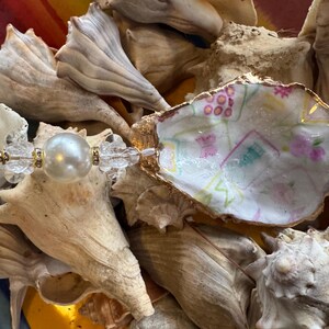 May include: A decorative oyster shell with a gold rim, hand-painted with a colorful design, and a pearl and crystal bead accent. The shell is surrounded by various seashells, creating a coastal theme.