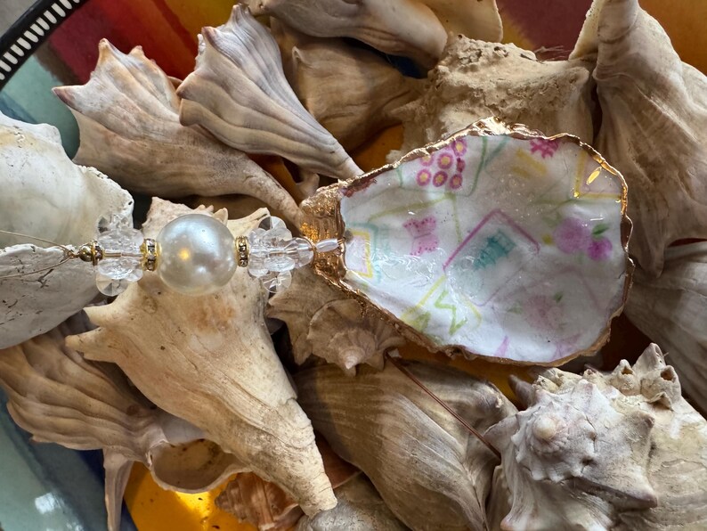 May include: A decorative oyster shell with a gold rim, hand-painted with a colorful design, and a pearl and crystal bead accent. The shell is surrounded by various seashells, creating a coastal theme.