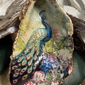 May include: A decorative oyster shell featuring a decoupage image of a peacock with vibrant blue, green, and gold plumage standing amidst colorful flowers. The edges of the shell are gilded with a metallic gold finish, creating a textured border.