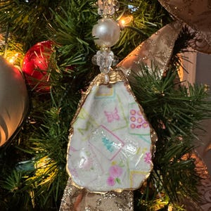 May include: A handmade Christmas tree ornament. The ornament is made from an oyster shell with a gold-painted edge. The shell is filled with a patterned fabric with pink flowers and butterflies. The ornament is topped with a pearl and crystal bead.