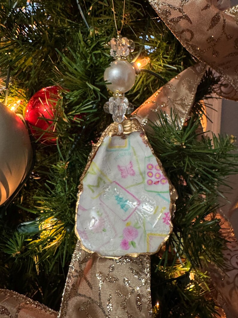 May include: A handmade Christmas tree ornament. The ornament is made from an oyster shell with a gold-painted edge. The shell is filled with a patterned fabric with pink flowers and butterflies. The ornament is topped with a pearl and crystal bead.