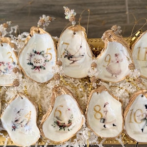 May include: A collection of twelve oyster shell ornaments, each with a gold-painted edge and a unique design. The ornaments feature numbers, floral patterns, and illustrations of figures in various poses. They are arranged in a gold tray.