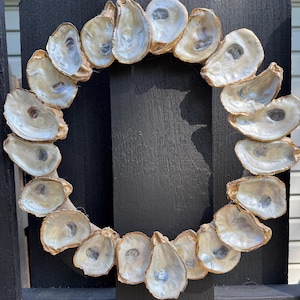 May include: A circular wreath made of oyster shells, with a natural, pearlescent interior and gold-painted edges. The wreath is suspended by a natural twine loop, and is displayed against a black wooden fence.