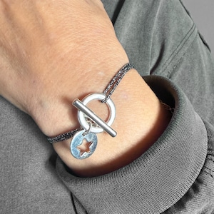 Peut inclure: Un bracelet composé d'un cordon texturé gris foncé et d'un fermoir à bascule argenté. Une breloque circulaire argentée avec une découpe en forme d'étoile est suspendue à l'anneau du fermoir. Le cordon se compose de deux brins fins.